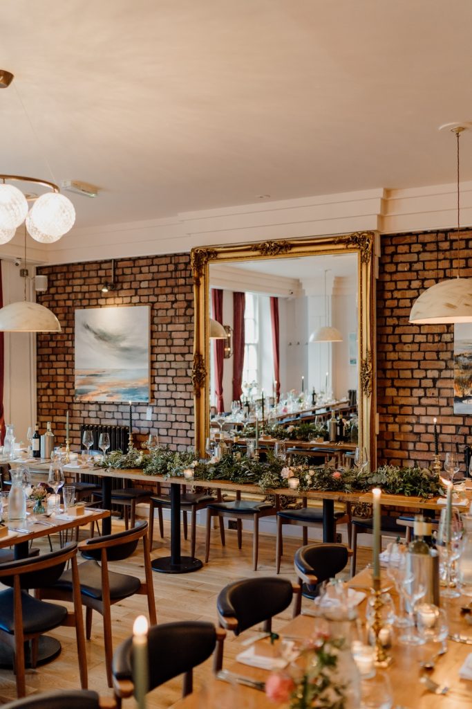 An elegantly decorated dining room at The Square Club, a prominent wedding venue in Bristol, is ready for a wedding reception. The warm and intimate setting features long wooden tables adorned with greenery, candles, and glassware, while an ornate gold-framed mirror reflects the room's charm. The exposed brick walls and modern lighting add to the venue's unique ambiance, making it an ideal spot for an intimate and stylish Bristol wedding celebration.