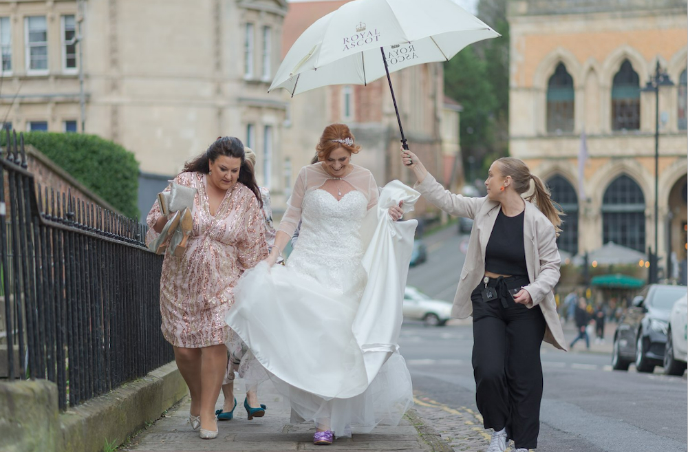 This image captures a candid moment before a wedding day at The Square Club, a wedding venue in Bristol. The bride, elegantly dressed in white, is walking alongside her bridesmaids while the in-house wedding coordinator holds an umbrella to shield her from the rain. There is a sense of excitement as the bride smiles and laughs.The historic streets of Bristol add a charming urban backdrop, reflecting the versatility of The Square Club, which blends contemporary city elements with timeless wedding traditions. This Bristol wedding venue provides the perfect setting for such joyful and spontaneous moments.