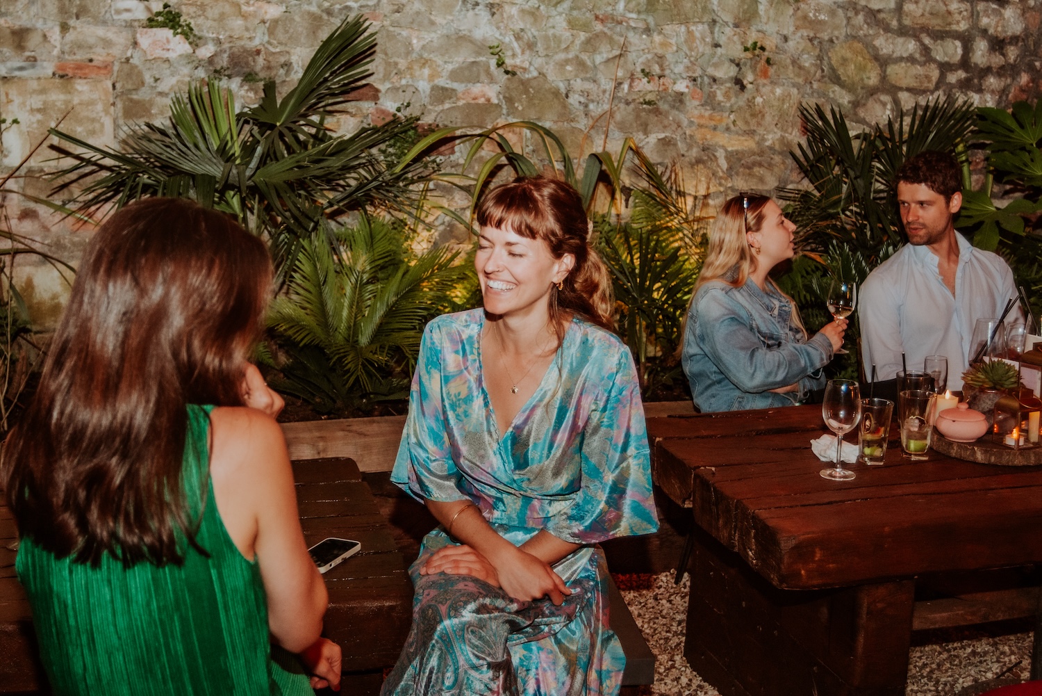 Two friends enjoying a relaxed conversation in the garden space of The Square Club, surrounded by tropical plants and rustic wooden tables. This serene setting highlights the venue's appeal as a unique and versatile events space in Bristol.