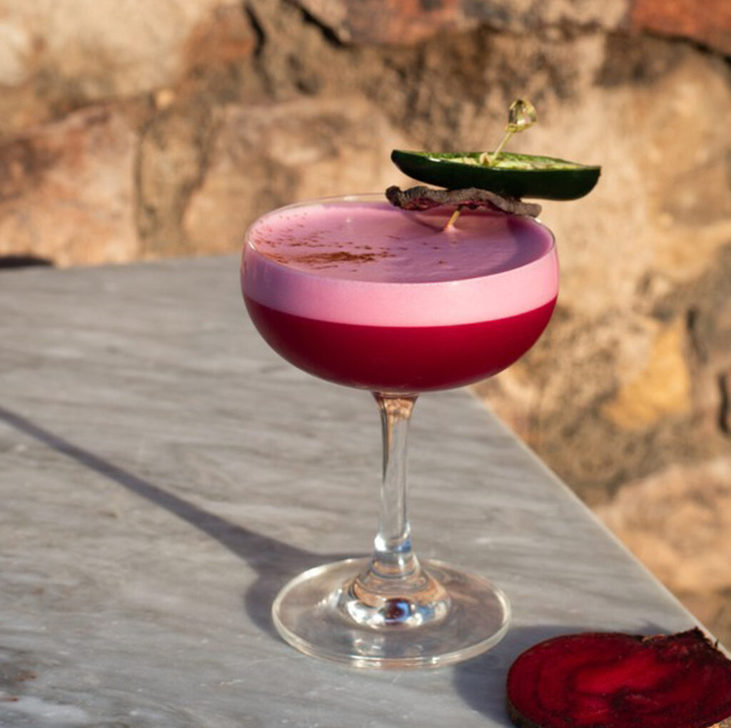 A vibrant pink cocktail garnished with a slice of cucumber and a delicate dusting of spices, served on a marble table at The Square Club. The rich colours and creative presentation highlight the exceptional craftsmanship of this Clifton restaurant in Bristol, making it a top choice for a sophisticated drink experience.