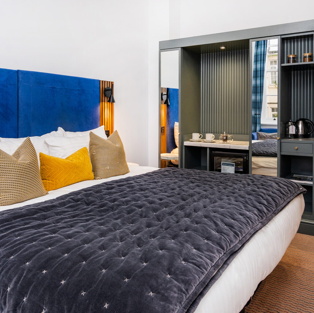 Contemporary bedroom at The Berkeley Suites, Bristol, part of The Square Club, featuring a deep blue upholstered headboard, cozy bedding, and a well-equipped workspace for both leisure and business travelers.