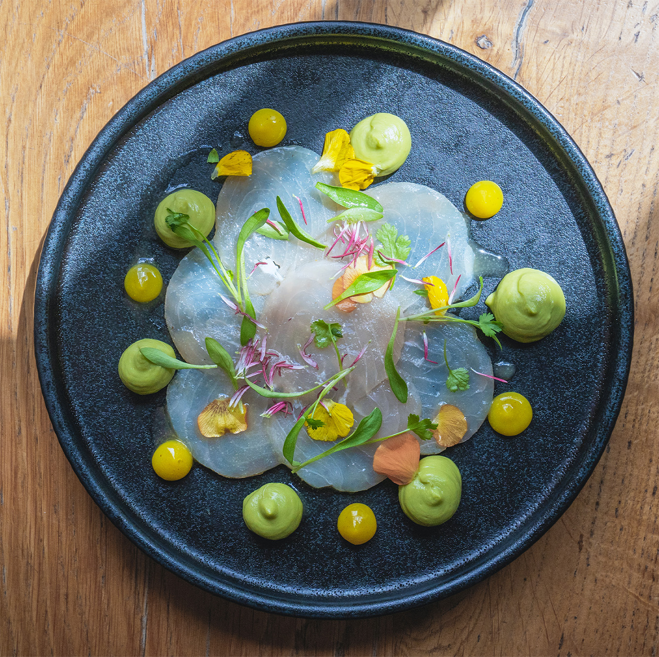 A delicate carpaccio of white fish, adorned with avocado purée, edible flowers, and citrus elements, on a black plate. This light and refreshing dish at The Square Club is a perfect choice for summer dining at one of the best restaurants in Bristol.