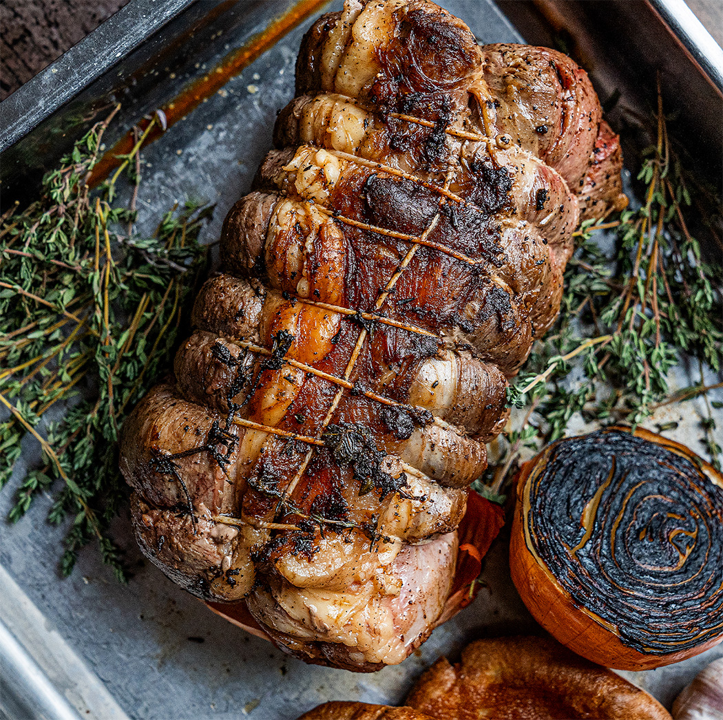 A perfectly roasted joint of beef, surrounded by charred onions, garlic, and sprigs of thyme, in a roasting tray. This classic dish from The Square Club demonstrates its culinary expertise and solidifies its reputation for offering the best Sunday lunch in Bristol within a cosy Clifton setting.