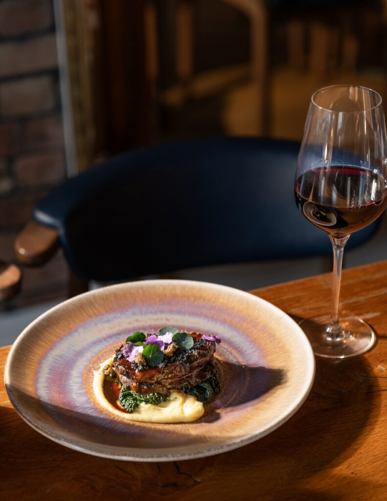 Beautifully plated lamb rump served on a bed of creamy mash with sautéed greens, garnished with edible flowers, paired with a glass of red wine at The Square Club restaurant in Bristol. A fine dining experience perfect for special occasions in Clifton.